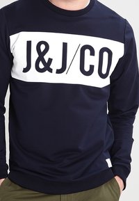 Mörkblå sweatshirt med en vit horisontell rand som har "J&J/CO" logotypen i fet svarta bokstäver. Ribbad halsringning och muddar, mjuk textur.