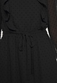 Robe noire avec de petits points texturés, manches longues, volants sur les côtés et une ceinture en tissu attachée à la taille.