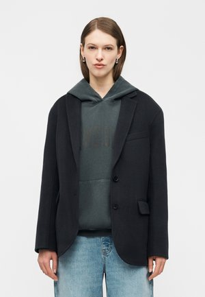 Žena s rovnými hnedými vlasmi nosí strieborné kruhové náušnice, tmavý nadrozmerný sako cez tmavozelenú mikinu s kapucňou a svetlomodré džínsy, smeruje dopredu.