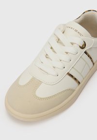 Vit lädersneaker med beige detaljer, rund tå och texturerad gummisula. Har snörning och geometriskt mönster på sidan.