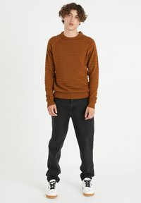 Suéter marrón de punto con rayas texturizadas, cuello redondo y puños acanalados, combinado con vaqueros negros y zapatillas blancas.
