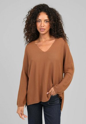 Femme aux cheveux bouclés portant un pull marron ample à col en V et un jean foncé, debout devant un fond gris uni.