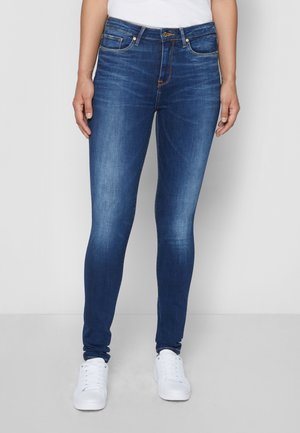 Jean bleu slim porté avec des baskets blanches et un haut blanc, montré de la taille aux pieds sur un fond uni.