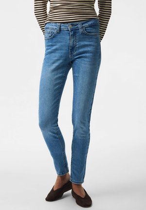 Person trägt hellblaue Skinny-Jeans, braune Slipper und ein langärmeliges schwarzes und beiges gestreiftes Oberteil vor einem einfarbigen Hintergrund.