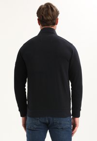 Hombre con cabello castaño claro, vistiendo una chaqueta negra de mangas largas y jeans azules, de pie de espaldas contra un fondo blanco.
