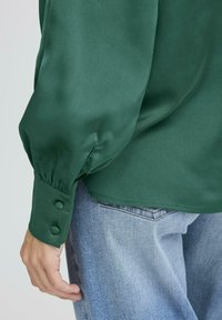 Blusa verde in satin con maniche a palloncino e polsini con bottoni, abbinata a jeans in denim azzurro chiaro. La texture sembra liscia e lucida.