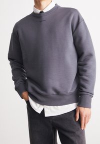 Felpa grigia in misto cotone con polsini e orlo a costine, presenta un colletto rotondo e il logo di Calvin Klein sul petto. Indossata sopra una camicia bianca.