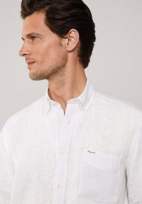 Camisa de lino blanca de botones con un bolsillo, que presenta una textura sutil, un collar clásico y un pequeño logo de la marca en el bolsillo.
