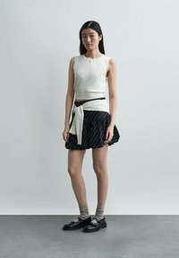 Fiatal nő hosszú sötét hajjal, ujjatlan fehér felsőt, fekete szoknyát fehér csíkkal, a derekán megkötött fehér pulóvert, szürke zoknit és fekete cipőt visel.