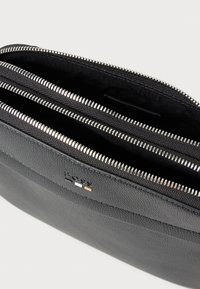 Černá kožená taška se třemi kapsami na zip, stříbrné kování a jemný strukturovaný povrch. Přítomen je detail s logem.