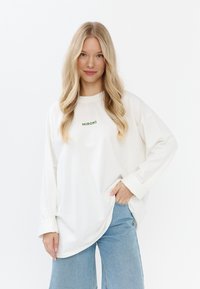 Übergroßer weißer Sweater mit grünem Text "MIRONS" auf der Brust, lange Ärmel und lockere Passform. Kombiniert mit Jeans.