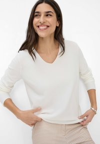 Weißer, gerippter Pullover mit langen Ärmeln und V-Ausschnitt. Hat eine lockere Passform und wird mit gemusterten beigen Hosen kombiniert. Glatte Textur, minimalistisches Design.