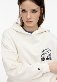 Bela kapuca s sproščenim krojem, ki ima na prsih grafiko žabe in besedilo "FROG MODE". Mehek material z rebrastimi manšetami in kapuco na vrvico.
