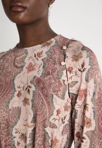 Blomsterdekket topp i myk rosa, med intrikat paisley-design, rund hals, og små knapper langs skuldersømmen.