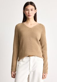 Tanfarget strikkegenser med V-hals, ribbestrikket tekstur og lange ermer; har skrå sømmer og en lett buet kant. Brukt med lyse bukser.