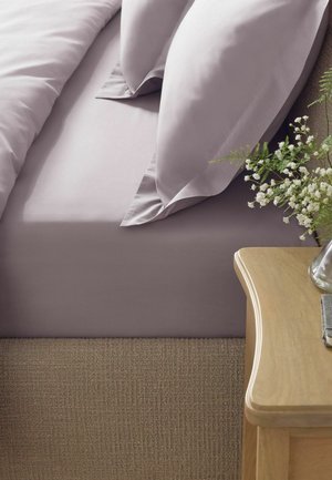 Lenzuola e cuscini viola su un letto con pavimento in moquette beige e un comodino in legno con un vaso di vetro con fiori bianchi.