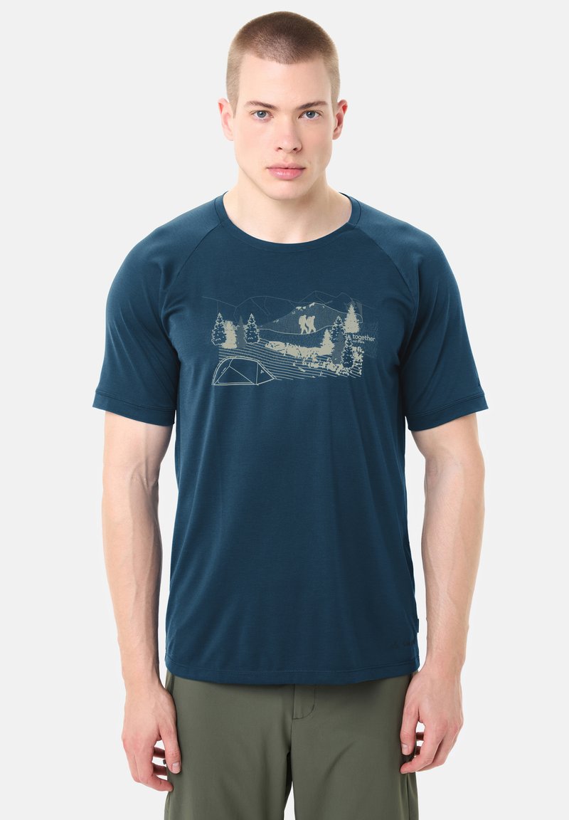 Jeune homme aux cheveux courts portant un t-shirt bleu marine avec un motif beige de randonnée et camping, associé à un pantalon vert olive.