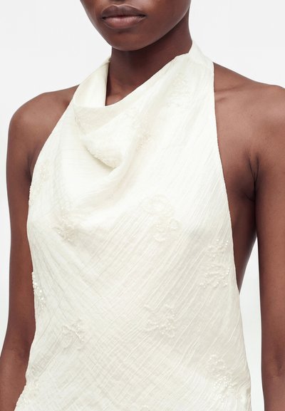 Γυναίκα που φοράει λευκό αμάνικο halter top με λεπτή κεντητή λουλουδάτη διακόσμηση και ντραπαρισμένο ντεκολτέ σε μονόχρωμο φόντο.
