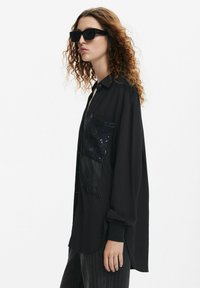 Camicia nera a maniche lunghe con tasca decorata da perline lucide e dettagli frangiati, vestibilità ampia, design con colletto, abbinata a occhiali da sole neri.