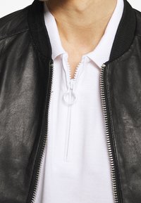 Man som bär en svart läderjacka över en vit zip-up polo tröja, som visar kragen och övre bröstområdet.