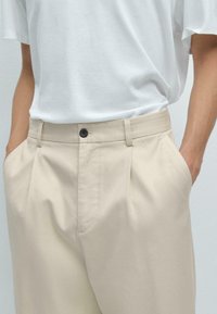 Beige pleated trousers with side pockets and a single black button closure at the waist, paired with a loose white T-shirt.