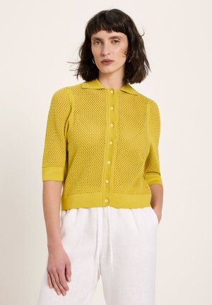 Frau mit kurzem dunklem Haar, die ein gelbes, kurzärmeliges, gestricktes Oberteil mit Knöpfen und weiße, hoch taillierte Hosen trägt, stehend vor einem schlichten Hintergrund.