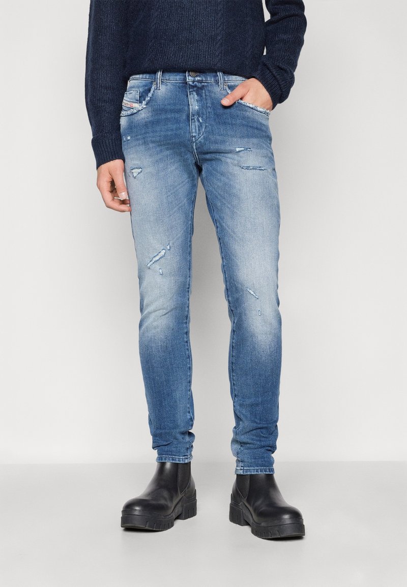 Personne vêtue d'un jean bleu délavé usé, de bottes noires épaisses et d'un pull bleu marine foncé à manches longues, debout avec une main dans la poche.