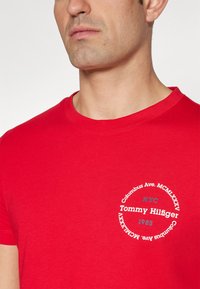 Czerwona bawełniana koszulka z okrągłym nadrukiem zawierającym napisy „Tommy Hilfiger”, „NYC” i „1985” w kontrastujących kolorach. Krótkie rękawy.