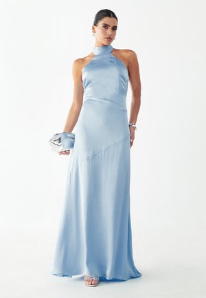 Kobieta w jasnoniebieskiej sukni na szyję bez rękawów, trzymająca srebrną kopertówkę, z srebrną bransoletką i sandałami na obcasie na białym tle.