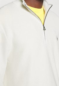 Weißer Halbzip-Pullover aus strukturiertem Stoff mit kontrastierender gelber Innenschicht. Verfügt über ein kleines Logo auf der Brust und einen Metallreißverschluss.