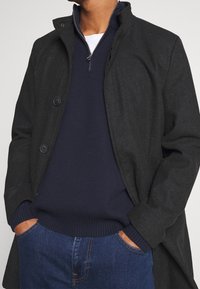 Cappotto in lana grigio scuro con collo alto, dotato di chiusura con bottoni. Indossato sopra un maglione blu navy con zip e jeans di denim.