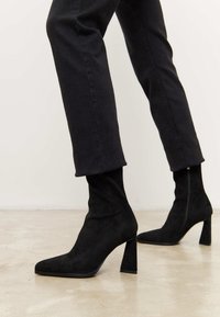 Botines de ante negro con punta afilada, tacón alto y cuadrado, y caña ajustada. Detalle de cremallera en el lateral.