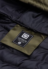 Interno di una giacca nera trapuntata con un'etichetta verde oliva, che presenta la dicitura "ombre menswear" e la taglia "M" stampate in bianco.