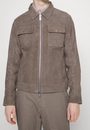 Veste en suede marron avec un col, fermeture éclair argentée et deuxpoches poitrine. Tissu texturé avec une coupe ajustée, associée à un pantalon à carreaux.