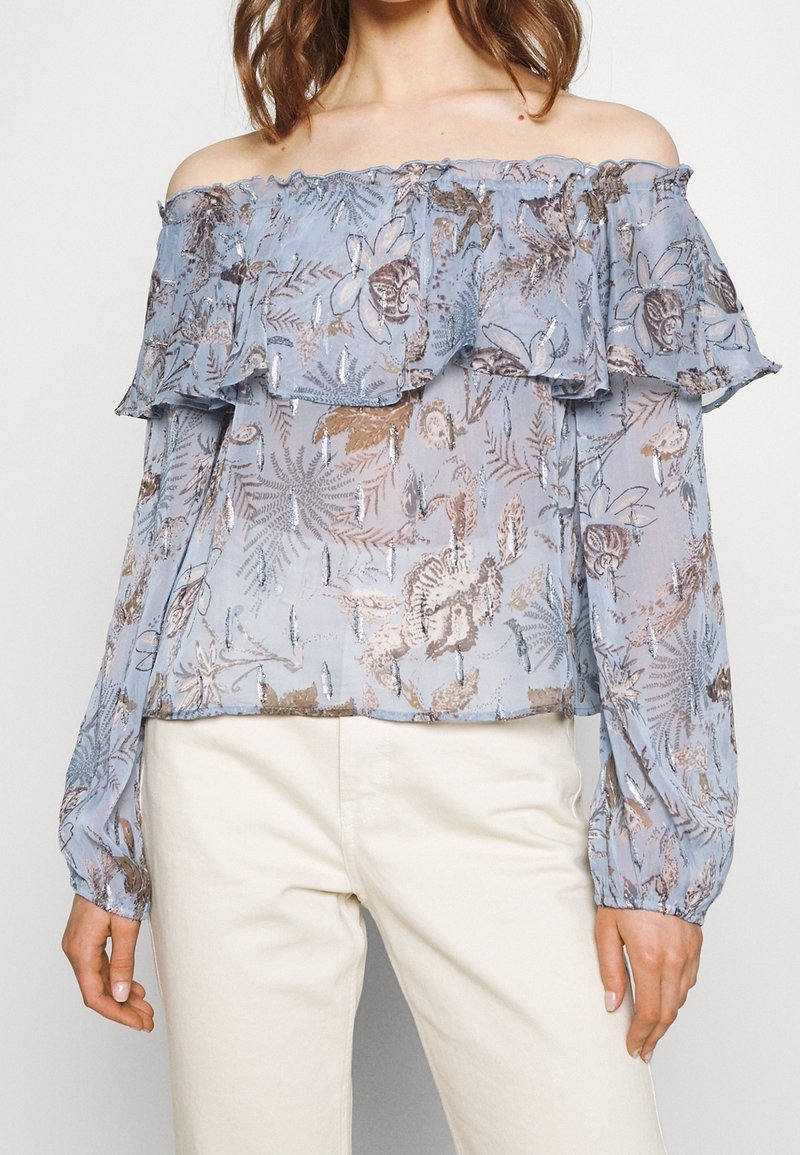 Blouse légère bleu clair transparente à épaules dénudées avec motifs floraux et feuilles, col volanté et longues manches bouffantes, portée avec un pantalon clair.