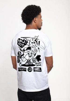 Uomo visto di profilo che indossa una maglietta bianca con un personaggio da cartone animato a tema basket in stile graffiti nero e la scritta "Who Not?" sul retro.