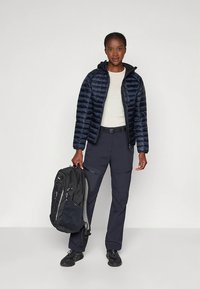 Tamsiai mėlyna pūkinė striukė su užtrauktuku, baltas viršus, tamsios darbo kelnės ir juodas kuprinė. Lygi tekstūra ir lieknas siluetas. Juodi sportbačiai užbaigia aprangą.