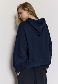 Tamnoplava oversized majica s kapuljačom na vezanje, rebrastim manžetama i rubom. Mekani materijal, opušteni kroj i minimalistički dizajn.