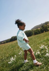 Niño pequeño con cabello rizado vestido con ropa azul claro camina por un campo de hierba con flores blancas bajo un cielo despejado.