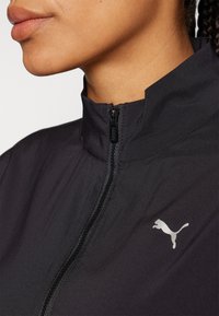 Chaqueta negra con cremallera y cuello alto, que presenta un logo plateado de Puma en el lado izquierdo y una textura de tela suave y ligera.