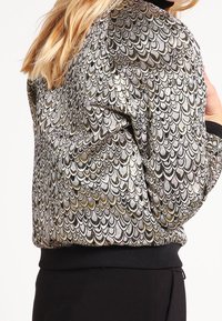 Metaliczna kurtka bomberka z wzorem łusek ryb w kolorze srebrnym, czarnym i złotym. Posiada prążkowane mankiety i dół, fakturowaną tkaninę oraz luźny krój.