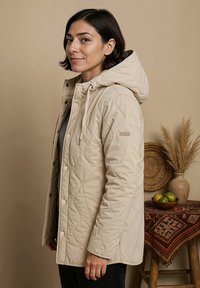 Beige gewatteerde jas met een capuchon, voorzien van drukknoopsluiting, trekkoorden en zijzakken, tegen een neutrale achtergrond met decoratieve items.