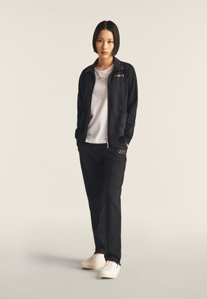 Tuta nera con giacca con zip e pantaloni abbinati, entrambi con dettagli dorati. Camicia bianca sotto, abbinata a sneakers bianche.