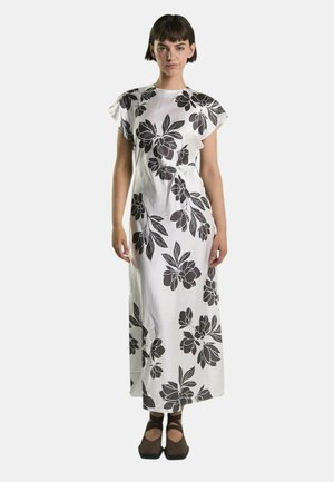 Donna in piedi che indossa un vestito maxi bianco con grandi stampe floreali nere e scarpe chiuse marroni con cinturini alla caviglia.