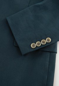 Manche de costume vert foncé comportant quatre boutons beige alignés près du poignet, avec des boutonnières visibles et une texture fine du tissu.