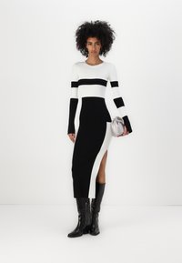 Lång klänning med svart och vit randig design, åtsittande passform, sprund på sidan och långa ärmar; kombinerad med knähöga svarta stövlar och en silverfärgad clutchtaska.