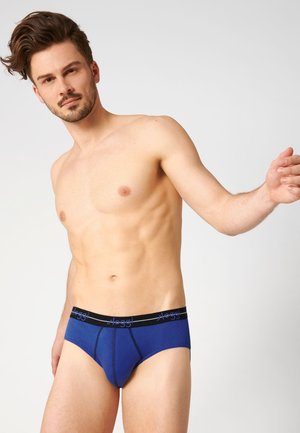 Hemdloser Mann, der blaue Unterhosen mit schwarzem Bund trägt, steht vor einem einfarbigen hellen Hintergrund, linker Arm nach außen gestreckt.