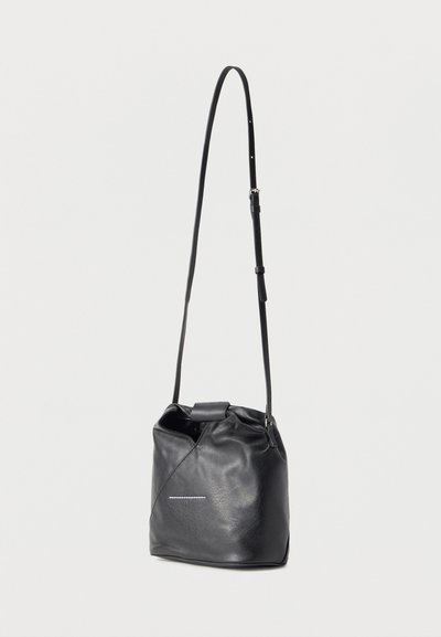 Μαύρη δερμάτινη τσάντα τύπου κάδος με ρυθμιζόμενο λουράκι ώμου και μικρή λευκή ραφή στο μπροστινό μέρος, παρουσιάζεται με φόντο ένα απλό υπόβαθρο.