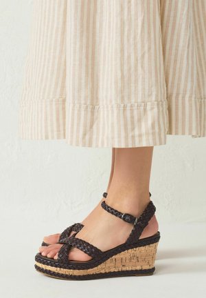 Voeten met donkerbruine gevlochten sleehakken met enkelbandjes, gecombineerd met beige en wit gestreepte wijde broekspijpen.