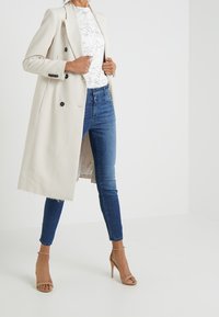 Ljusbeige dubbelknäppt kappa med svarta knappar, lager-på-lager över en vit linjetecknad grafisk skjorta och mörkblå smala jeans. Ljusbruna remsandaler.